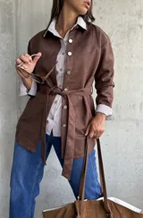 Sobrecamisa de gamuza color chocolate, con cierre frontal de botones metálicos y lazo ajustable en la cintura. Presenta cuello clásico y mangas largas con puños rectos.