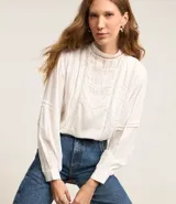 Blusa blanca de viscosa con cuello redondo, manga larga con puños ajustados y detalles de entremedios huecos y frizos delanteros.