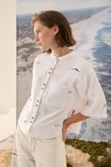 Camisa de lino blanca con cuello mao, mangas cortas y cierre frontal de botones. Presenta delicados bordados multicolores distribuidos por la prenda y bolsillos frontales en el pecho.