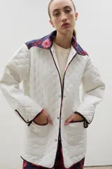 Campera acolchada de corte holgado, color blanco con diseño capitoneado en forma de rombos. Presenta cuello camisero y bordes internos con estampado floral en tonos azul y rojo, cierre frontal con botones a presión y bolsillos laterales.