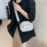 Cartera bandolera color gris claro, de cuero sintético, con correa ancha de tela estampada y cierre superior con cremallera.