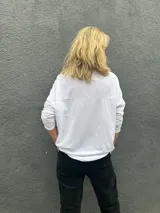 Blusa blanca de corte holgado y diseño minimalista, con cuello redondo y mangas largas. Presenta una textura sutilmente arrugada en la tela.