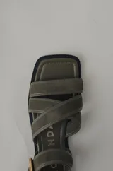 Sandalia plana de cuero negro, con diseño minimalista de tiras finas cruzadas sobre el empeine y una tira ajustable con hebilla metálica en el tobillo.