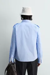 Camisa de algodón color celeste, corte relaxed fit, con cuello solapa, manga larga y trabillas en los hombros. Presenta dos bolsillos de plastrón con solapa en el pecho.