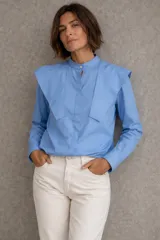 Camisa de popelina de algodón en color azul, con cuello mao y solapas estructuradas en los hombros que aportan volumen. Presenta cierre frontal oculto con botones y mangas largas con puños clásicos.