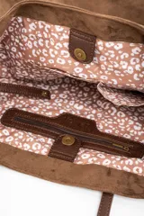 Bolso tipo tote de gran tamaño confeccionado en gamuza sintética color tostado. Presenta un diseño minimalista con asas largas de hombro y una pequeña placa metálica frontal con el logo de la marca.