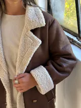 Tapado de estilo aviador en color chocolate con interior y detalles de corderito en color beige. Presenta cierre frontal cruzado con botones, solapas amplias y puños con borde de corderito.