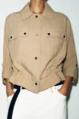 Chaqueta corta marrón de cuello solapa y manga remangada por debajo del codo. Presenta bolsillos delanteros de plastrón con solapa, bajo ajustable con cordón interior y cierre frontal con botones.