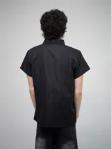Camisa de manga corta color negro, con corte regular y cierre frontal de botones. Presenta un estampado gráfico en blanco en el lado derecho del pecho con motivos artísticos y tipografía gótica.
