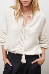 Camisa de color crudo con diseño romántico, cuello redondo con cordón ajustable terminado en borlas, y mangas tres cuartos abullonadas. Presenta detalles de bordados calados verticales en el frente y horizontales en las mangas.