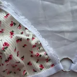 Conjunto de lencería de dos piezas confeccionado en tela acanalada blanca con estampado de pequeñas flores rosas. Incluye un soutien tipo triángulo sin aro, con detalles de encaje blanco en los bordes y una argolla metálica en el centro, y una bombacha colaless ajustable con tiras laterales y detalles de encaje.