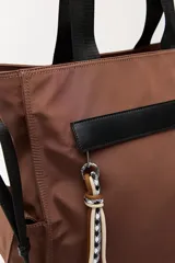 Bolso tipo tote color negro, con doble asa superior y correa desmontable. Presenta un bolsillo frontal con solapa, bolsillos laterales y un detalle colgante decorativo con tiras de colores.