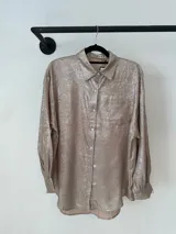 Camisa de corte holgado en tono beige metalizado, con cuello clásico, cierre frontal de botones, bolsillo en el pecho y mangas largas con puños abotonados.