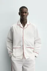 Pijama de dos piezas confeccionado en popelín de algodón blanco, con vivos de ribete en color bordó. Incluye una camisa de manga larga con cuello bowling, bolsillos de plastrón y cierre frontal de botones, y un pantalón de corte relajado con cintura elástica y bolsillos laterales.