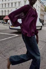 Blusa de tejido semitransparente color violeta, con cuello redondo y detalle de fornitura dorada en el escote. Presenta un diseño de escote en pico, mangas largas abullonadas con puños elásticos y corte holgado.