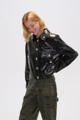 Chaqueta corta de eco-cuero negra con cuello camisero, cierre frontal con botones plateados y bolsillos simulados con botones plateados.