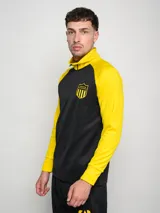 Buzo deportivo de entrenamiento, color negro en el torso y amarillo en las mangas y cuello. Es de manga larga, con cuello alto y cierre parcial frontal. Presenta un escudo con estrellas en el pecho.