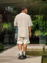 Camisa de manga corta color crudo/blanco, confeccionada en tejido de punto abierto con textura calada y cuello polo. Presenta cierre frontal de botones.