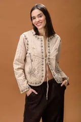 Chaqueta acolchada de color beige con bordados lineales y florales en hilo negro. Presenta un diseño de corte recto, manga larga y cierre frontal mediante lazos finos.