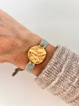 Pulsera con dije redondo dorado con diseño abstracto y cinta de terciopelo elástico color gris claro.