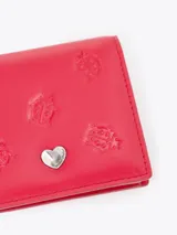 Mini billetera de cuero vacuno color rojo, con diseño grabado de pequeñas mariquitas en el frente y un aplique metálico en forma de corazón. Cuenta con un interior funcional con espacios organizadores para tarjetas.