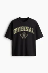 Remera negra de punto pesado de algodón con cuello redondo, hombros caídos y diseño holgado. Presenta un estampado frontal estilo universitario con la palabra "ORIGINAL" en tono desgastado y un gráfico circular rodeado de laureles.