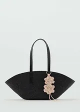 Bolso tipo capazo de fibra natural en color negro, con forma de media luna. Cuenta con dos asas largas de hombro en color negro y un detalle decorativo de flores en tonos claros colgando de una de las asas.