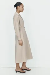 Vestido midi de color beige con cuello solapa y escote en pico. Presenta mangas largas, cintura elástica en la espalda y un cinturón a contraste con hebilla metálica. Incluye bolsillos laterales ocultos.
