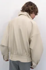 Chaqueta bomber oversize de efecto piel en color crema, con cuello subido, manga larga con hombro caído y cintura con frunces y cinturón ajustable con hebilla metálica. Cuenta con cierre frontal oculto y bolsillos laterales de vivo.