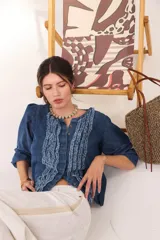 Camisa de jean de algodón liviano con escote redondo, cierre frontal con botones, mangas tres cuartos abullonadas y pechera con apliques de tiras deshilachadas.
