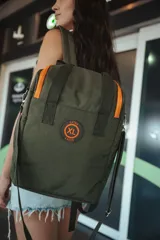 Bolso verde con detalles en naranja, con doble asa de mano, correa bandolera desmontable y correas traseras para usar como mochila. Tiene cierre doble, dos bolsillos externos con cierre, un bolsillo interno con cierre y dos bolsillos internos abiertos. Compartimentos internos para notebook y matero. Logo "XL Extra Large" estampado en naranja.