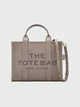 Cartera tipo tote de cuero granulado color tostado, con diseño estructurado y rectangular. Presenta doble asa superior fija y una correa larga ajustable y extraíble. En el frente, destaca el logo estampado con la inscripción 'THE TOTE BAG MARC JACOBS'.