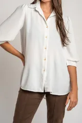 Blusa de corte oversize en color blanco, con cuello camisero, cierre frontal de botones y mangas tres cuartos con sutiles frunces en los hombros.