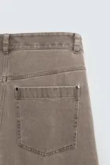 Pantalón de corte relajado en color marrón con efecto lavado. Presenta bolsillos frontales tipo cargo, cierre con cremallera y botón, y una línea horizontal de tachas metálicas a tono en la parte superior de las piernas.