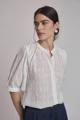 Camisa blanca con estampado de cruces, cuello mao y mangas 3/4 con detalle de pliegues en los hombros. Cierre frontal con botones.