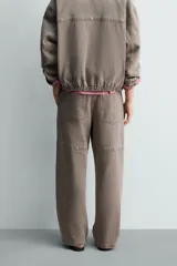Pantalón de corte relajado en color marrón con efecto lavado. Presenta bolsillos frontales tipo cargo, cierre con cremallera y botón, y una línea horizontal de tachas metálicas a tono en la parte superior de las piernas.