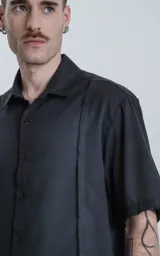 Camisa de manga corta en color negro con proceso de lavado, cuello clásico y cierre frontal con botones. Presenta un diseño minimalista con una costura vertical decorativa en el frente.