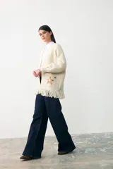 Chaqueta de punto color crema, con diseño abierto, mangas largas y flecos en el ruedo. Presenta un aplique bordado en forma de estrella en uno de los bolsillos frontales.