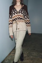 Chaqueta de punto con escote en pico y manga larga, confeccionada en tejido jacquard con diseño geométrico. Presenta detalles de lentejuelas y abalorios, cierre frontal con botones y un cinturón fino ajustable en la cintura.