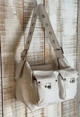 Bolso de hombro color beige con textura de cuero grabado tipo cocodrilo. Presenta dos bolsillos frontales con solapa y cierres metálicos, y una correa ancha ajustable con ojales metálicos.