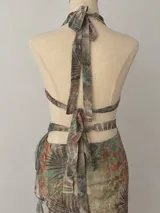 Vestido midi con estampado botánico en tonos tierra y verdes, con escote halter profundo y detalle de argolla metálica dorada en el centro. Presenta aberturas laterales en la cintura y falda con volados laterales.