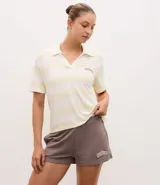 Remera polo deportiva de manga corta, confeccionada en tejido piquet con textura. Presenta un diseño a rayas horizontales en tonos crema y blanco, con cuello tipo polo y bordado con la palabra 'Athletics' en el pecho.
