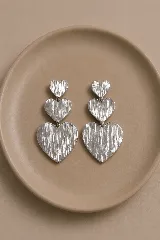 Aros colgantes plateados compuestos por tres corazones de tamaño decreciente con textura rugosa y acabado metálico.