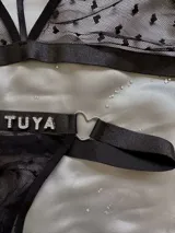 Conjunto de lencería de dos piezas en tul negro con estampado de corazones floca, compuesto por un bralette con tiras ajustables y argolla en forma de corazón, y una tanga con tiras regulables, argollas metálicas y detalle de pedrería con la inscripción 'Soy Tuya'.