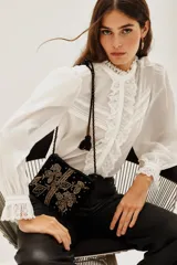 Camisa blanca de cuello alto con volados y encaje, mangas largas abullonadas con puños de encaje. Presenta detalles de encaje y pliegues verticales en el frente.