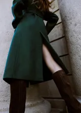 Abrigo largo de paño en color verde oscuro, con cuello alto, cierre frontal oculto y cinturón ajustable del mismo tejido. Presenta un diseño estructurado con bolsillos laterales.