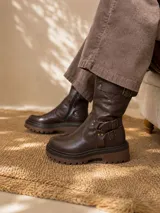 Botas de caña media confeccionadas en cuero color marrón oscuro, con suela track de goma en tono marrón más claro. Presentan un diseño con múltiples correas y hebillas metálicas decorativas en la parte inferior y superior de la caña, además de un cierre lateral interno.