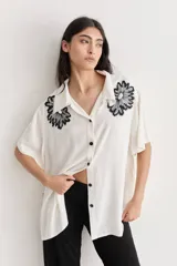 Camisa color crudo de corte amplio con bordados florales negros en la parte superior. Tiene cuello camisero, mangas cortas y cierre frontal con botones negros.