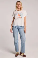 Remera blanca de algodón y poliéster, con cuello redondo y diseño holgado. Cuenta con un estampado frontal de inspiración vintage con la frase "Spritzing the day away", un fondo de tablero a cuadros en tonos celestes y la ilustración de una copa de spritz junto a naranjas y hojas.