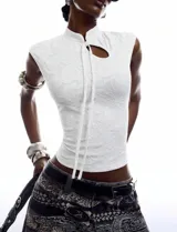 Top blanco de encaje floral con diseño de inspiración oriental, corte al cuerpo, cuello alto, abertura frontal con lazo para anudar y mangas cortas.